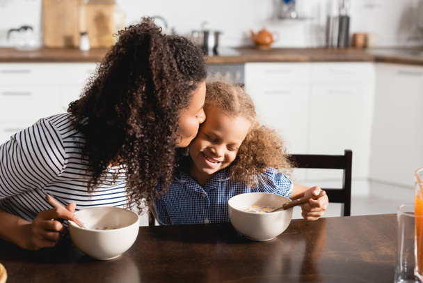young african american mother kissing daughter during breakfast in kitchen - Foto, immagini