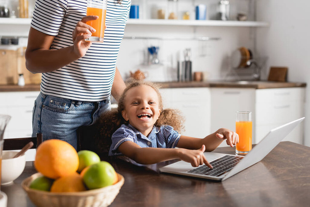 cropped view of woman with glass of orange juice near excited african american daughter using laptop in kitchen - Fotoğraf, Görsel