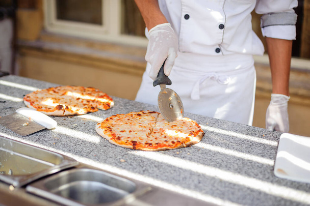 Hand van chef-kok bakker in wit uniform snijden pizza - Foto, afbeelding
