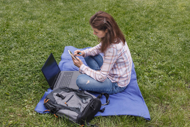 Werkplaats op een blauw tapijt in de natuur. Vrouw in de tuin naast de laptop met een stift in haar hand en kijkend naar de telefoon - Foto, afbeelding