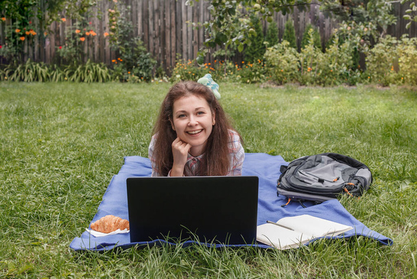 Workplace on a blue rug in nature. Woman lying on her stomach smiling near laptop and looking at camera. Next to her is a backpack, a notebook and a croissant - Photo, Image