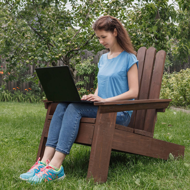Young woman in jeans and a blue T-shirt remotely working on a laptop sitting on a chair in the garden - Photo, Image