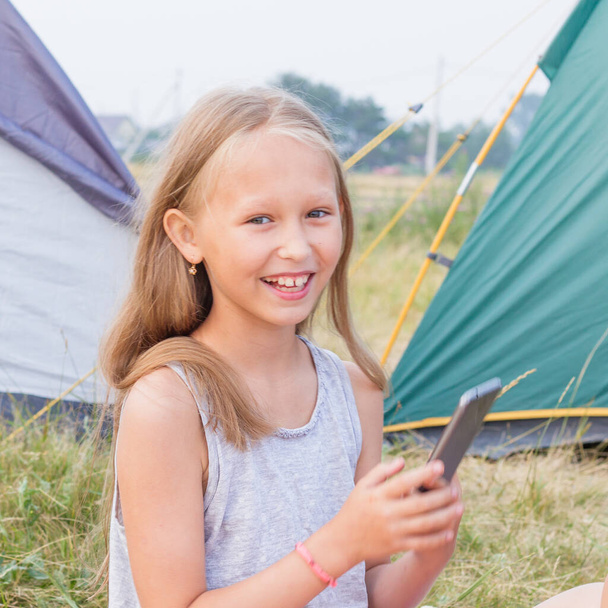 Cute little girl with long blond hair takes a picture on a smartphone and smile on a background of tents. - Photo, Image