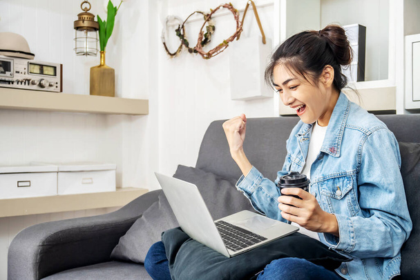 Asyalı genç bir kadın evdeki bir kanepede oturup kahve içerken bilgisayarını kullanarak internet üzerinden ve evden para kazanıyor. - Fotoğraf, Görsel