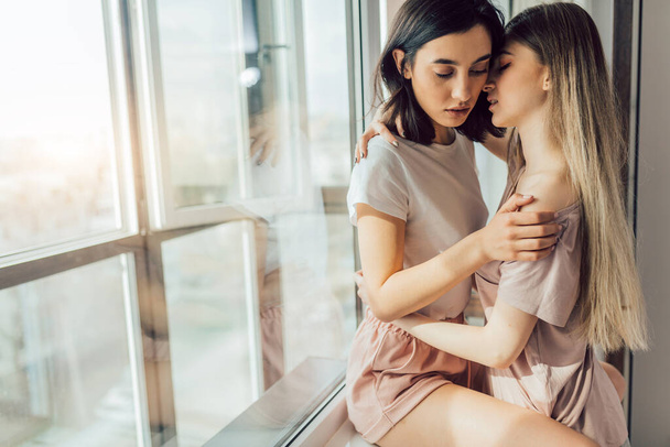 Deux lesbiennes embrassant ensemble tout en se tenant près de la fenêtre dans la chambre. - Photo, image
