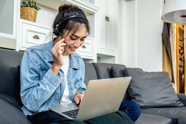 アジアの若いビジネスの女性は彼女自身の家のソファの上に座っているマイクと自宅のノートパソコンとオンラインビデオ会議で話している、自宅からの仕事 - 写真・画像