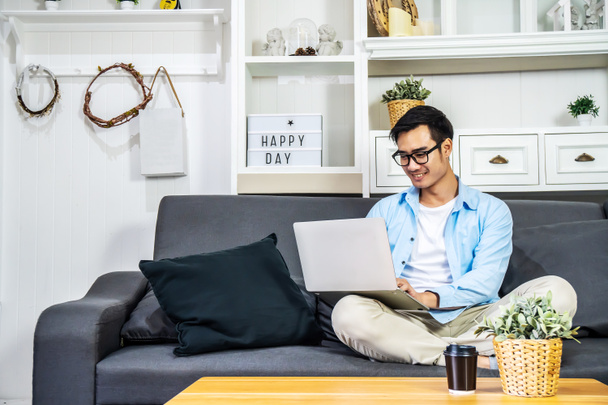Smart asiatischen Geschäftsmann in lässiger Kleidung sitzt auf einem Sofa im Haus genießt die Arbeit mit dem Computer-Laptop, um Geld aus dem Online-Geschäft zu verdienen, arbeiten von zu Hause aus - Foto, Bild