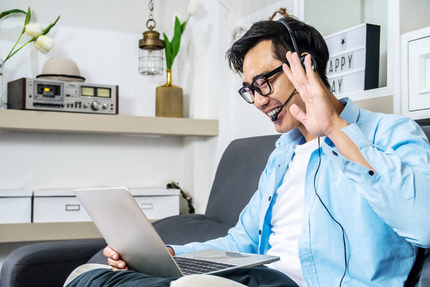Ázsiai fiatal üzletember ül a kanapén a saját házában visel fejhallgató videó hívás konferencia laptop számítógép emelje fel a kezét azt mondja hello csapat otthon iroda, munka otthonról - Fotó, kép
