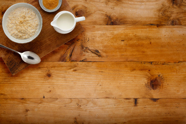Een voedzame kom pap, een kleine kom bruine suiker en een kan melk met een lepel, op een houten plank, op een houten achtergrond met kopieerruimte - Foto, afbeelding