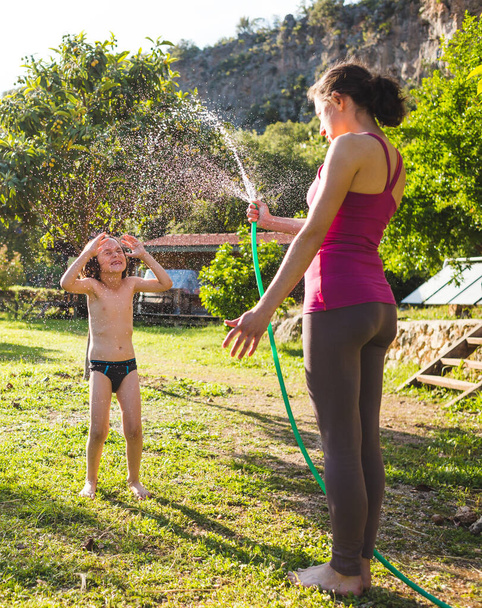 Η μητέρα ψεκάζει ένα παιδί με ένα σωλήνα στην αυλή του σπιτιού, αγόρι μούσκεμα στο νερό σε μια ζεστή ηλιόλουστη μέρα, Ψυχαγωγία στις διακοπές, Τα συναισθήματα ενός παιδιού, Παιδική διασκέδαση. - Φωτογραφία, εικόνα