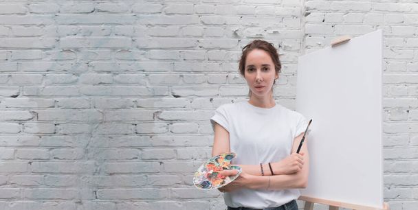 Beautiful woman artist posing at camera, brick wall background, free space for copy text, white t-shirt, brush in hand, palette for paints. White canvas, easel. - Photo, image