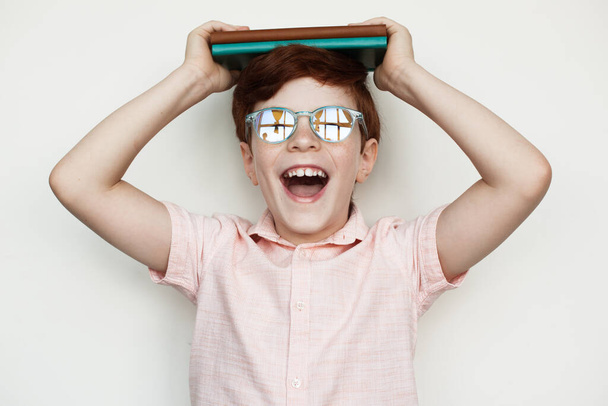 Jonge blanke jongen met gember haar met wat boeken op zijn hoofd en glimlach naar de camera met een bril op. - Foto, afbeelding