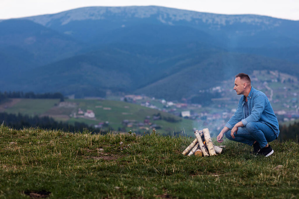 Χαρούμενος τουρίστας κάθεται δίπλα στη φωτιά, ταξιδιώτης χαμογελώντας στον καπνό της φωτιάς. τουρίστας καπνίζει μια φωτιά στο βουνό. - Φωτογραφία, εικόνα