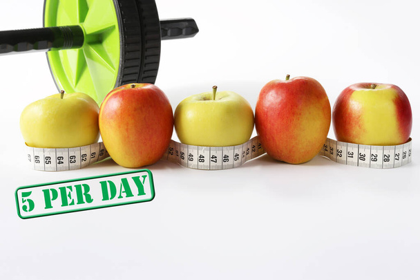 Healthy life concept. Multicolored apples with measure tap and ab wheel on white background - Photo, Image