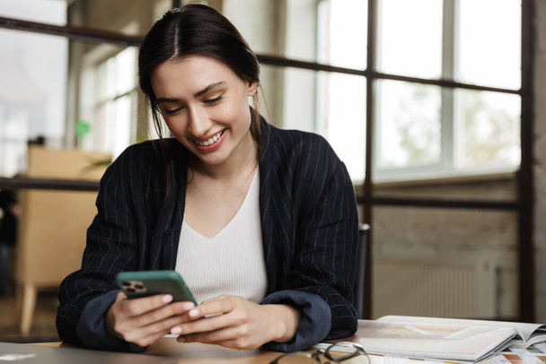 Confident smiling attractive young woman entrepreneur sitting at the office desk, using mobile phone - Foto, Imagen