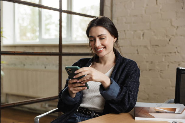 Confident smiling attractive young woman entrepreneur sitting at the office desk, using mobile phone - Fotó, kép