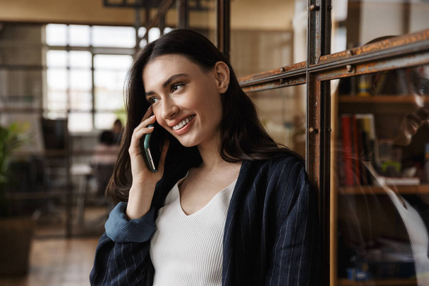 Aantrekkelijke glimlachende slimme jonge vrouw ondernemer staat terwijl leunend op een glazen muur, praten op mobiele telefoon - Foto, afbeelding