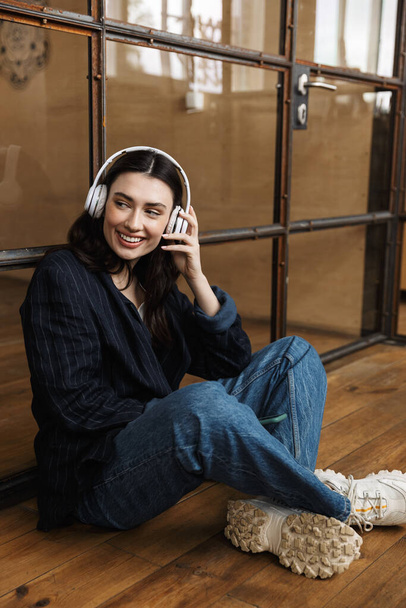 Attraktive, glückliche junge Frau sitzt im Büro, an eine Glaswand gelehnt, trägt kabellose Kopfhörer - Foto, Bild