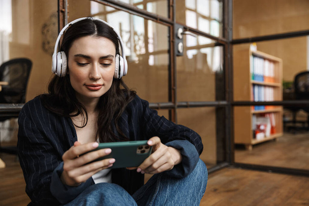 Attraktive junge Frau sitzt im Büro, lehnt an einer Glaswand, trägt kabellose Kopfhörer, benutzt Handy - Foto, Bild