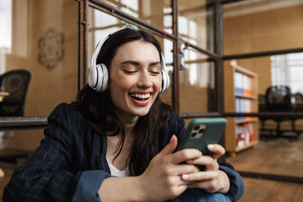 Attractive happy young woman sitting at the office, leaning on a glass wall, wearig wireless headphones, using mobile phone - Φωτογραφία, εικόνα