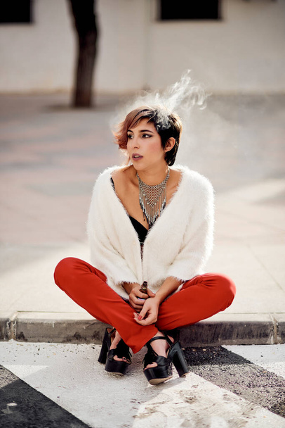 νεαρή κοπέλα που κάθεται στο πάτωμα όμορφα ντυμένη καπνίζοντας ένα ατμό με καπνό γύρω της - Φωτογραφία, εικόνα