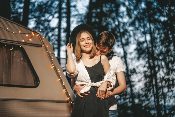 Young couple in love in stylish fashionable outfit romantic weekend wedding trip against background of camper in woods forest road trip - Fotó, kép