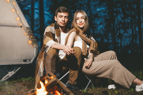 Νεαρό ζευγάρι στην αγάπη σε ζεστό και κομψό ντύσιμο κοντά στο τζάκι ρομαντικό Σαββατοκύριακο με φόντο το τροχόσπιτο στο δάσος οδικό ταξίδι - Φωτογραφία, εικόνα