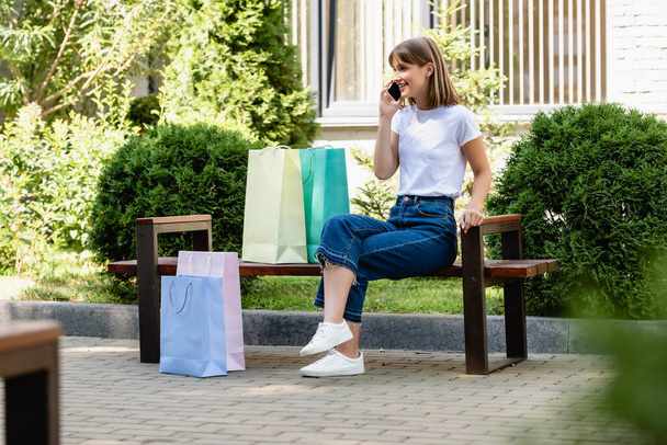 Selective focus of woman talking on smartphone near shopping bags on bench on urban street  - Foto, imagen
