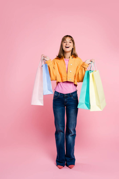 Excited woman in yellow jacket holding purchases and looking at camera on pink background - Photo, image