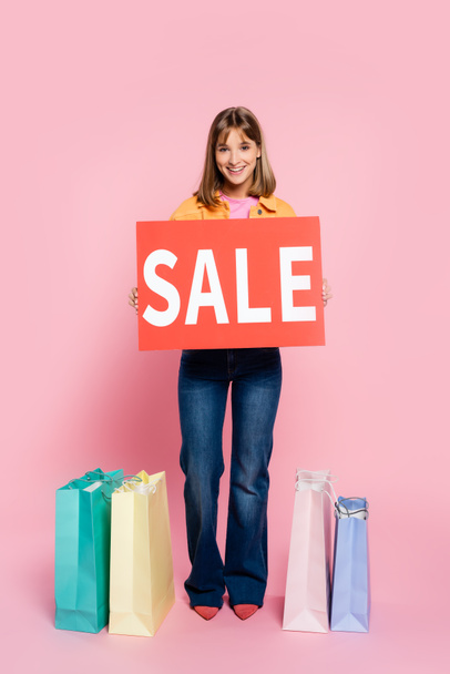 Young woman holding card with sale lettering near shopping bags on pink background - Photo, Image