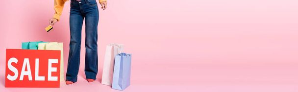 Panoramic crop of woman holding credit card near card with sale lettering and purchases on pink background - Foto, immagini