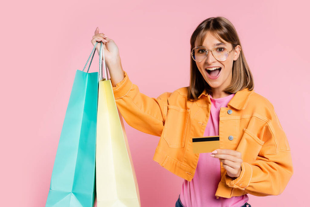 Excited woman in yellow jacket and sunglasses holding colorful shopping bags and credit card on pink background - Fotoğraf, Görsel