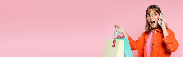 Panoráma lövés meglepett nő gazdaság bevásárló táskák árcédulák, miközben beszél okostelefon rózsaszín háttér - Fotó, kép