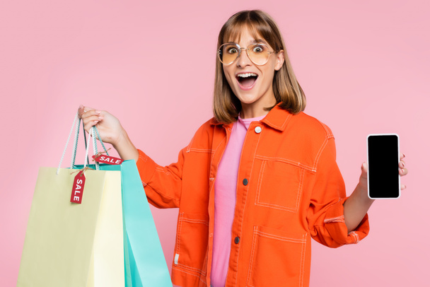 Mujer emocionada mostrando teléfono inteligente con pantalla en blanco mientras sostiene bolsas de compras con etiquetas de precios aislados en rosa  - Foto, imagen