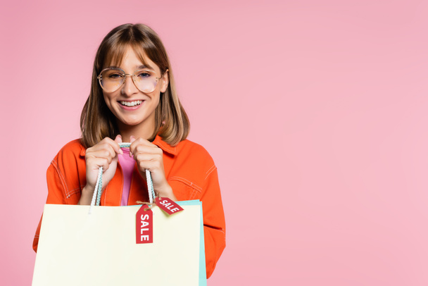 Young woman in sunglasses looking at camera and holding shopping bags with sale word on price tags isolated on pink  - Foto, Bild