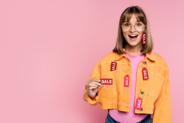 Excited woman showing price tag with sale word at camera on pink background - Фото, изображение