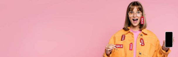 Изображение шокированной женщины с ценником и смартфоном на розовом фоне  - Фото, изображение