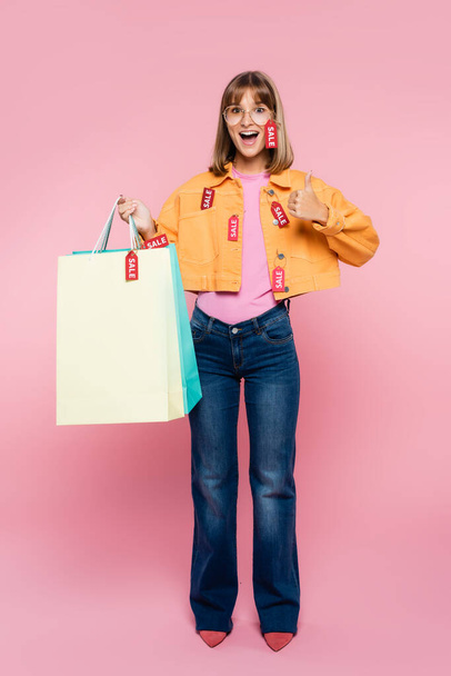 Excited woman holding shopping bags with price tags and showing thumb up on pink background - Photo, image