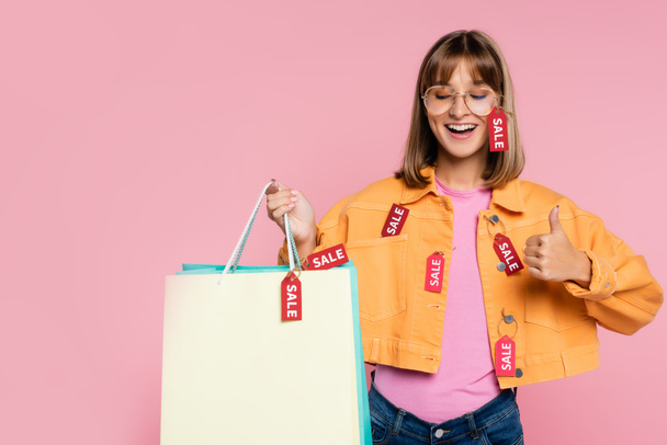Woman with closed eyes holding shopping bags with price tags and showing thumb up isolated on pink  - Photo, Image