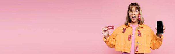 Panoramic shot of shocked woman in sunglasses showing price tag with sale word and smartphone with blank screen on pink background - Photo, image
