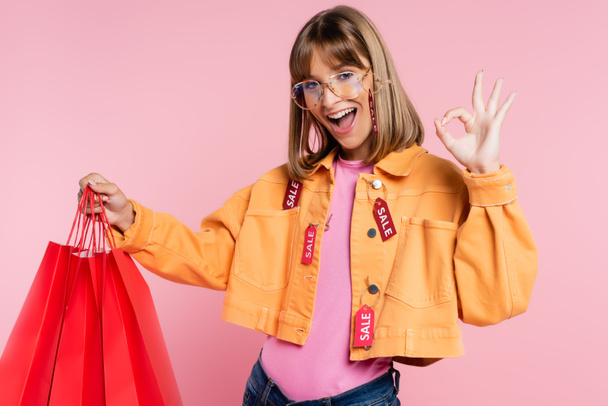 Woman with price tags on jacket holing red shopping bags and showing okay gesture on pink background - 写真・画像
