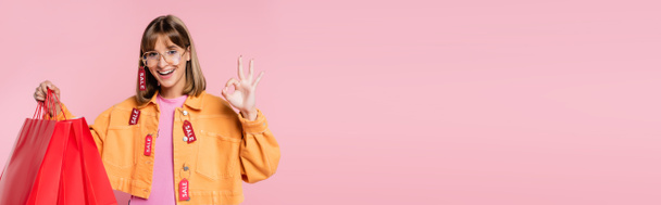 Panoramic concept of woman with price tags on jacket showing ok sign and holding red shopping bags isolated on pink  - Foto, Imagen