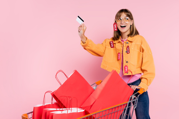 Excited woman with price tags on jacket holding credit card near shopping bags in cart on pink background - Фото, зображення