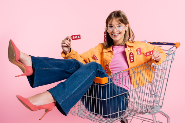 Szelektív fókusz a fiatal nő mutatja árcédulák eladó betűkkel kamera a bevásárlókocsi rózsaszín háttér - Fotó, kép