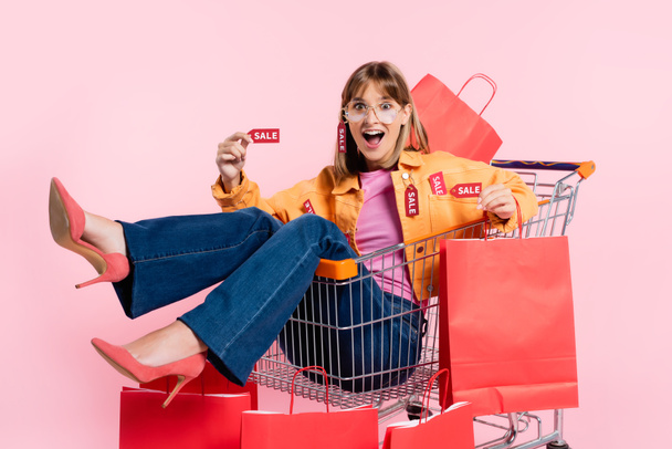 Mujer excitada en tacones y gafas de sol que muestra la etiqueta del precio y el bolso de la compra en el carrito sobre fondo rosa - Foto, imagen