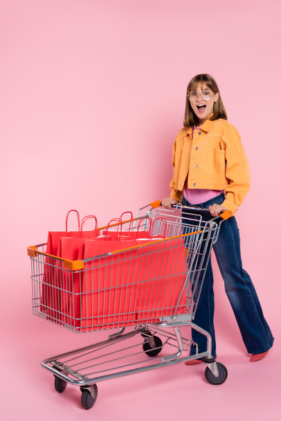Excited woman standing near red shopping bags in cart on pink background - Фото, зображення