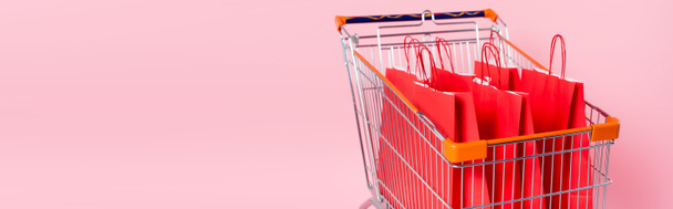 Panoramic shot of cart with red shopping bags on pink background - Photo, Image