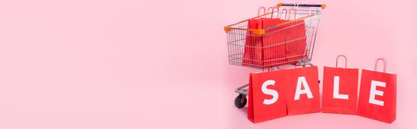 Conceito panorâmico de venda de palavras em sacos de compras vermelhos perto do carrinho na superfície rosa - Foto, Imagem
