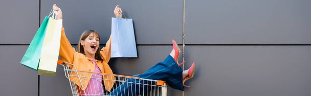 Horizontal image of excited woman holding colorful shopping bags while sitting in cart near building  - Fotografie, Obrázek