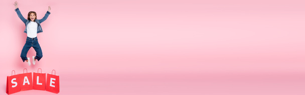 Imagen panorámica de mujer emocionada saltando cerca de bolsas de compras con letras de venta sobre fondo rosa - Foto, Imagen